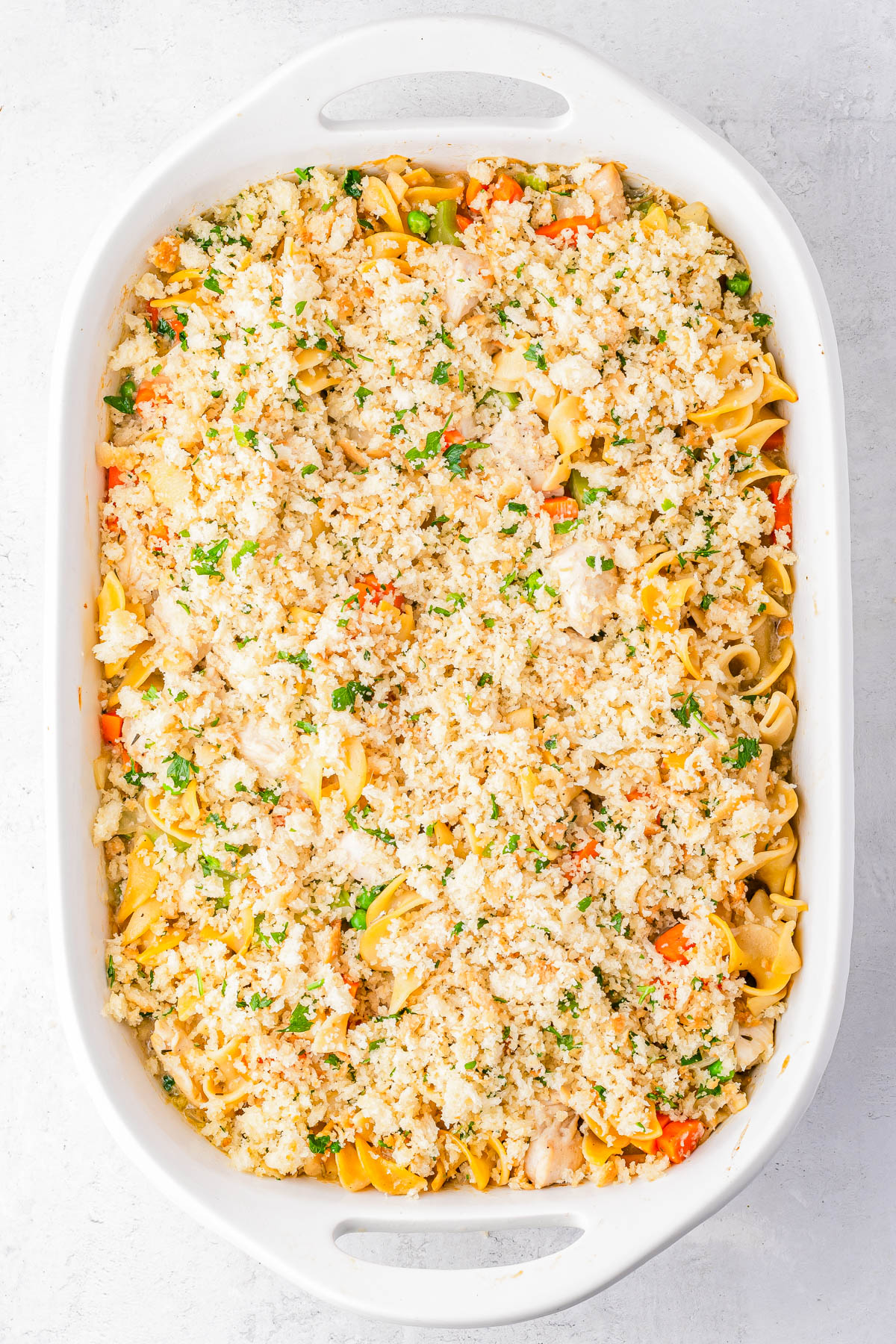 A white baking dish filled with a casserole topped with a crumbly mixture, containing visible pasta, vegetables, and herbs.