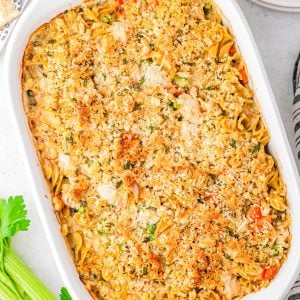 A baked casserole in a white dish topped with a golden breadcrumb crust, with vegetables and uncooked celery stalks nearby on a white surface.