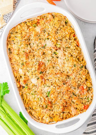 A baked casserole in a white dish topped with a golden breadcrumb crust, with vegetables and uncooked celery stalks nearby on a white surface.