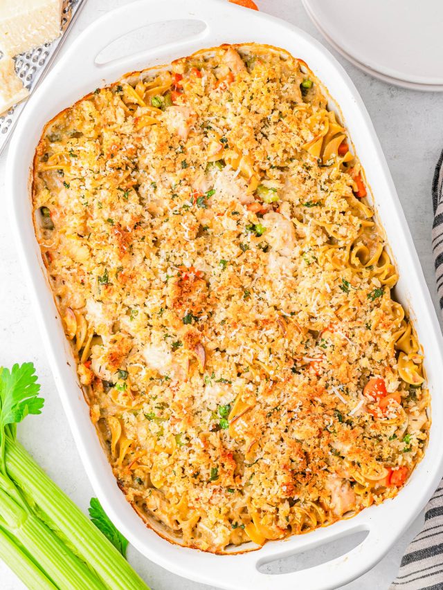 A baked casserole in a white dish topped with a golden breadcrumb crust, with vegetables and uncooked celery stalks nearby on a white surface.