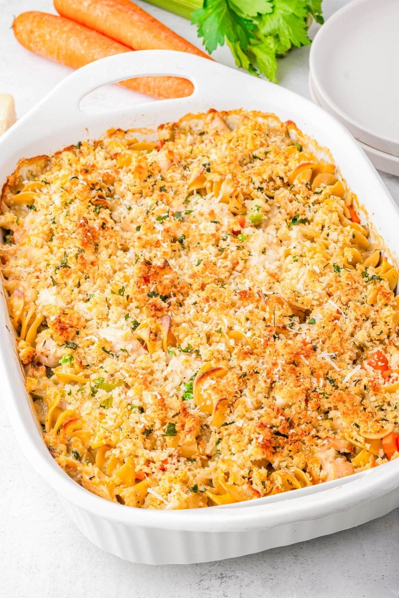 A baked casserole topped with breadcrumbs and herbs in a white dish, with visible noodles, vegetables, and pieces of meat. Plates, carrots, and celery are nearby.