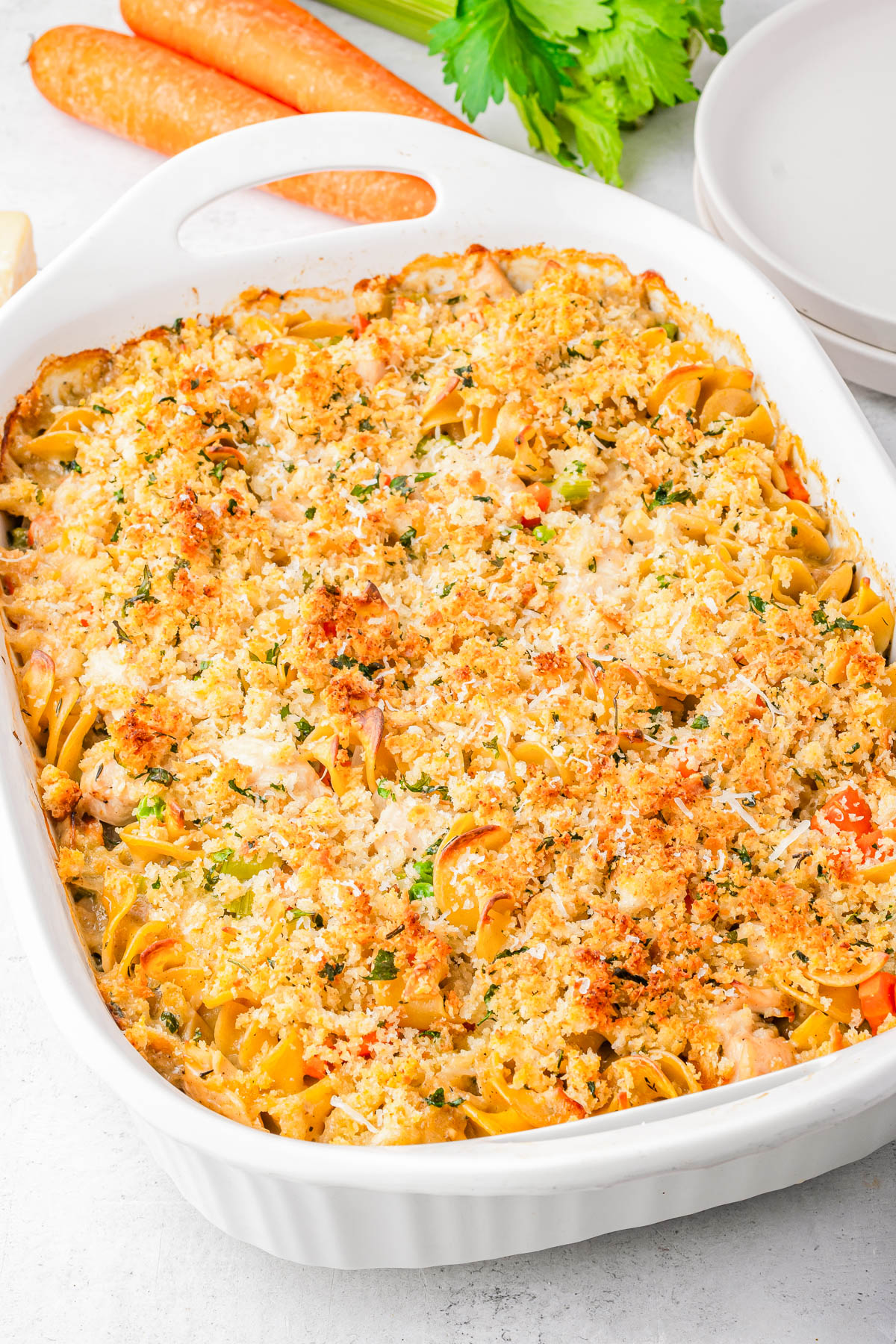A baked casserole topped with breadcrumbs and herbs in a white dish, with visible noodles, vegetables, and pieces of meat. Plates, carrots, and celery are nearby.