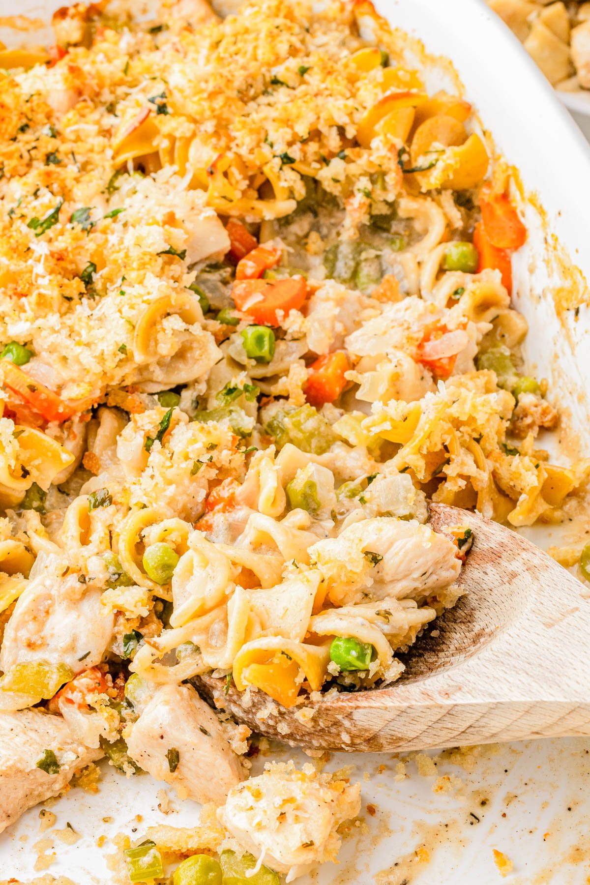 A close-up of a chicken noodle casserole with vegetables, topped with breadcrumbs, in a white baking dish with a wooden spoon scooping a portion.