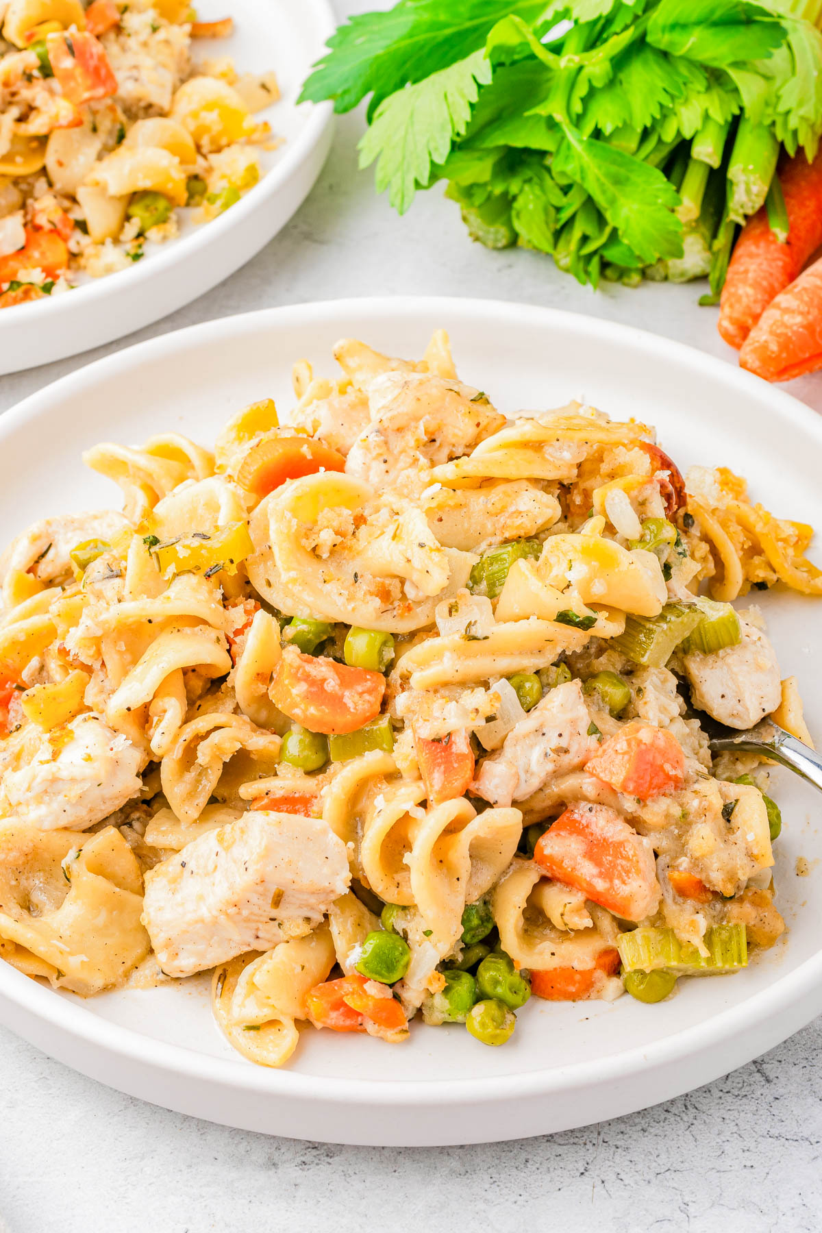 A plate of pasta mixed with chunks of chicken, carrots, peas, celery, and creamy sauce sits next to fresh celery and carrots.