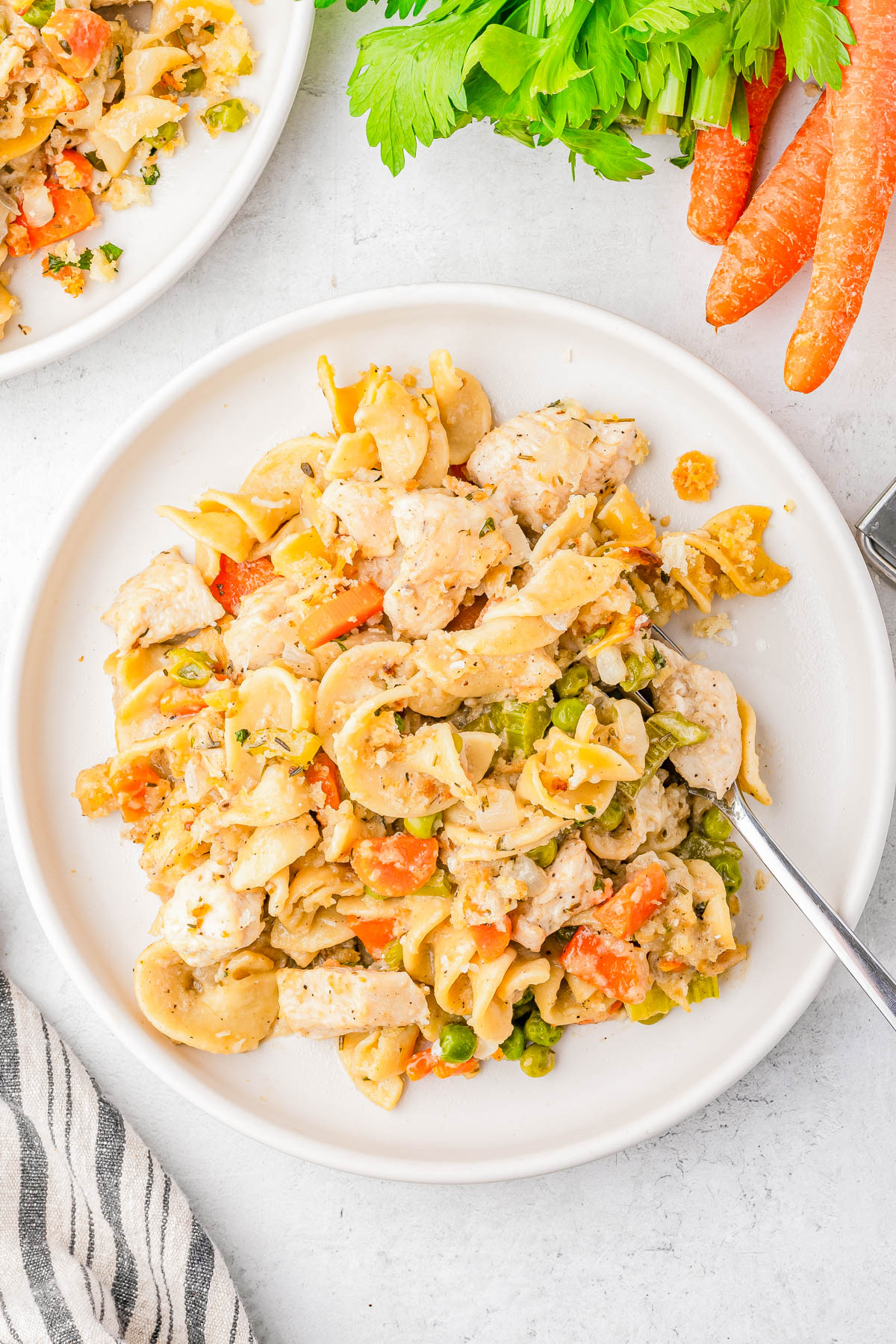 A plate of chicken noodle casserole with vegetables, including carrots and peas, next to fresh carrots and parsley on a white surface.