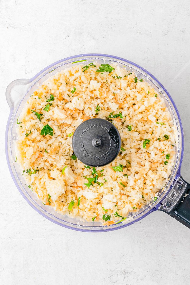 A food processor filled with finely chopped cauliflower mixed with green herbs, placed on a white surface.