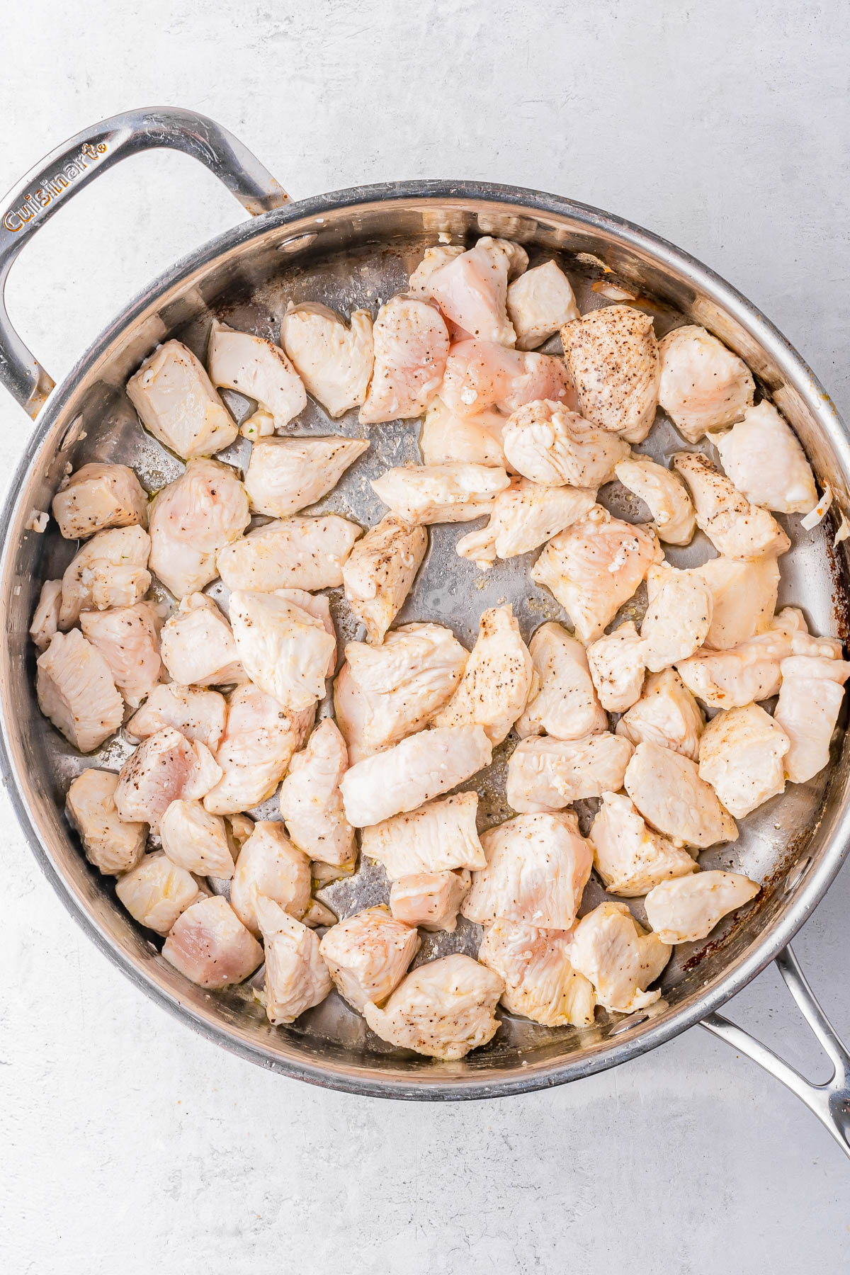 Diced raw chicken breast pieces seasoned with pepper cooking in a stainless steel skillet on a light surface.