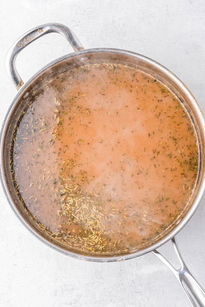 A stainless steel pot on a white surface containing simmering broth with herbs floating on top.