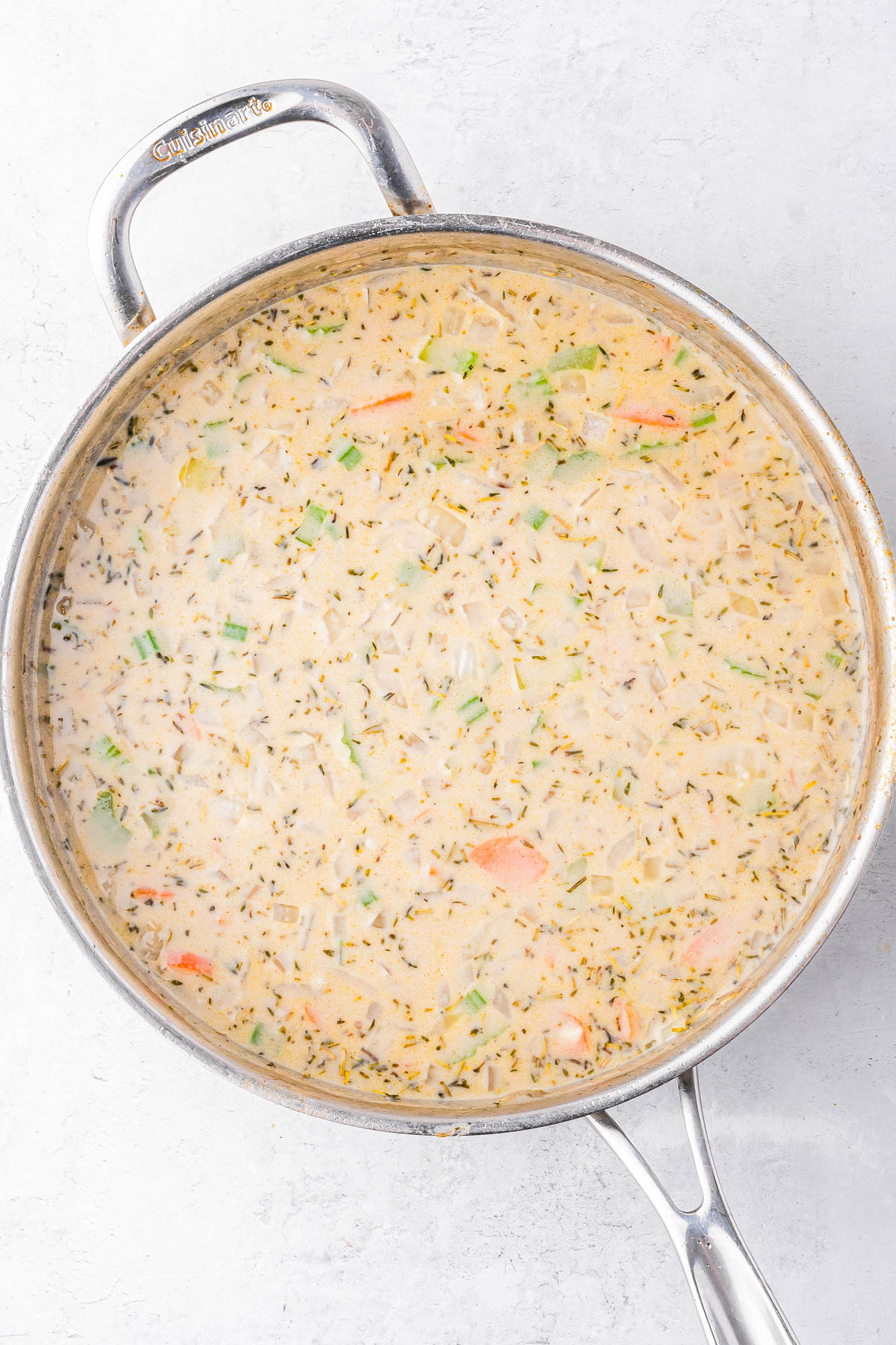 A stainless steel pan filled with creamy soup containing herbs, diced vegetables, and shredded chicken, viewed from above on a light surface.