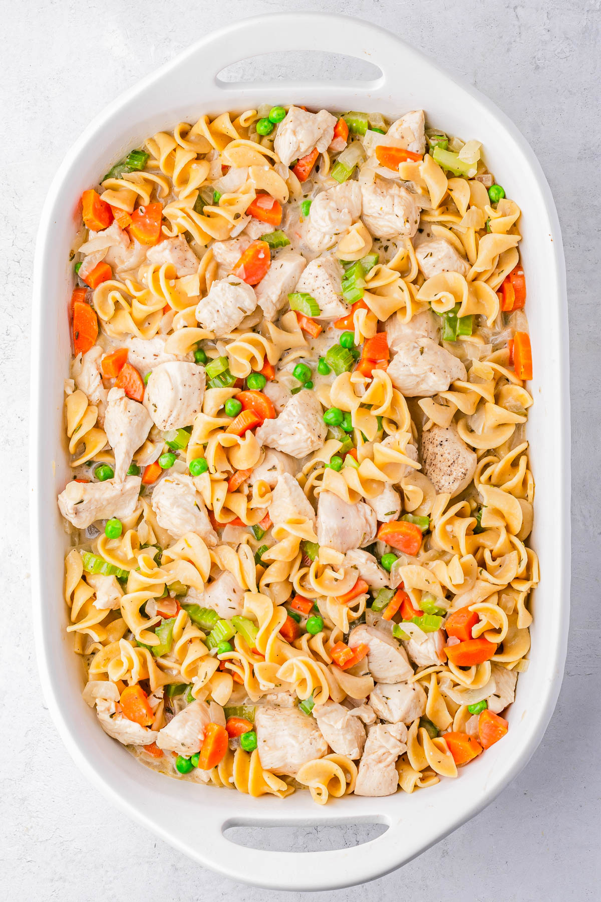 A white baking dish filled with cooked egg noodles, chunks of chicken, sliced carrots, peas, and celery.