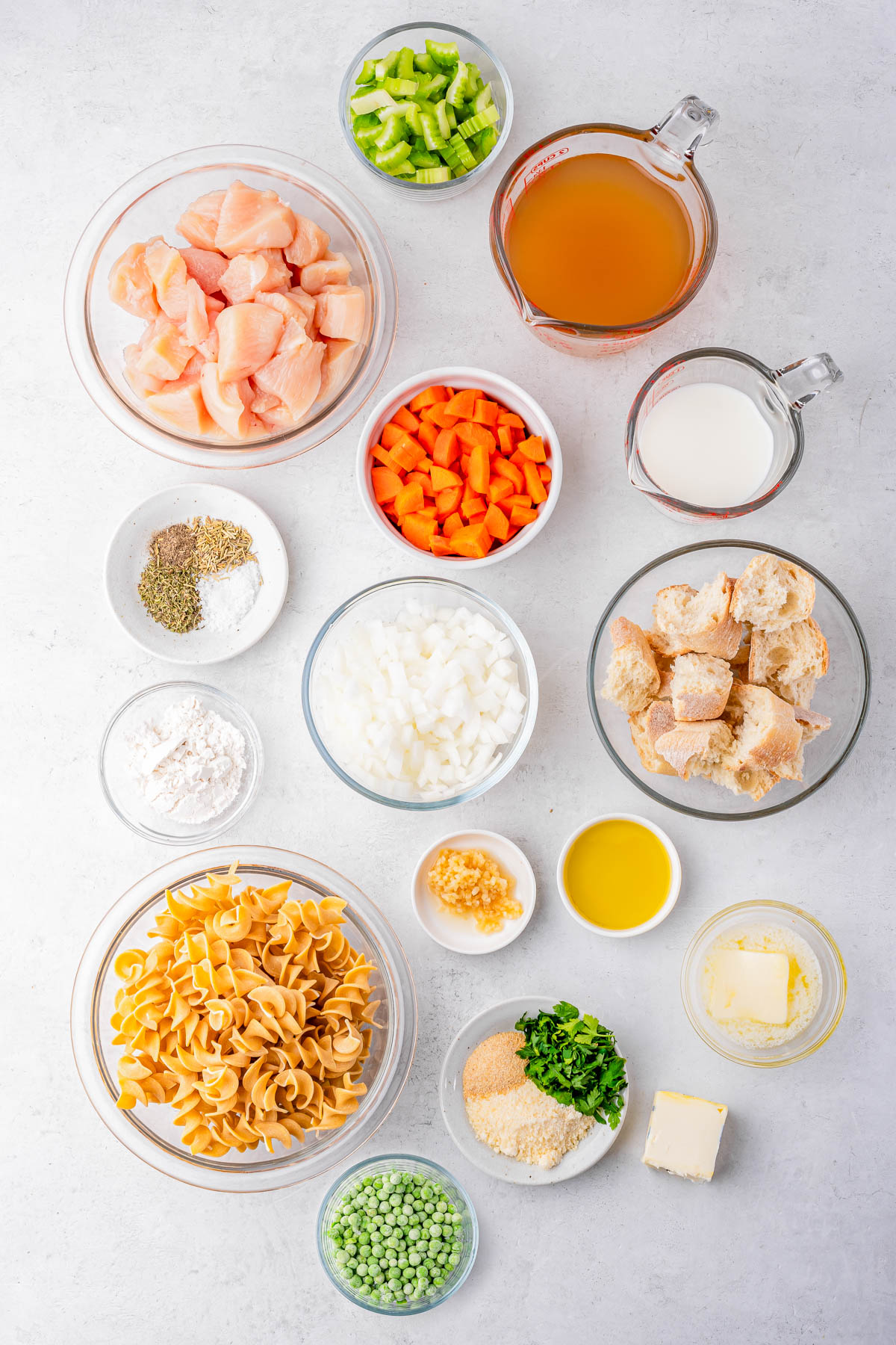 Various ingredients are arranged in bowls on a white surface, including cubed chicken, chopped vegetables, dry pasta, bread cubes, peas, seasonings, broth, milk, oil, and butter.