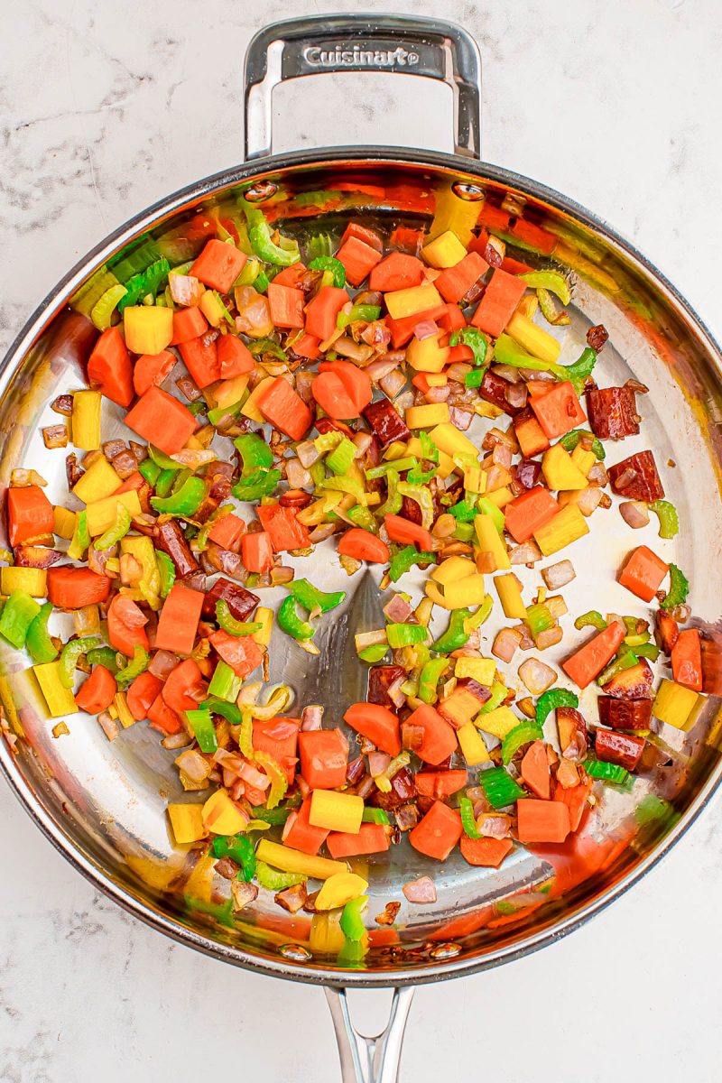 Diced red, yellow, and green bell peppers, onions, and sausage are sautéing in a stainless steel skillet on a marble countertop.