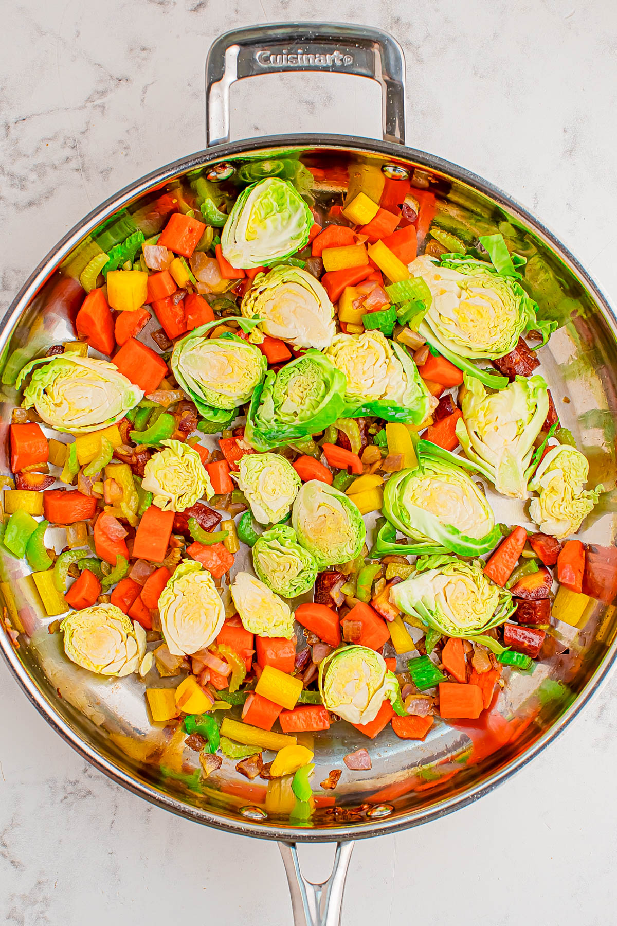 A stainless steel skillet filled with halved Brussels sprouts, chopped bell peppers, carrots, and onions, all arranged on a marble countertop.