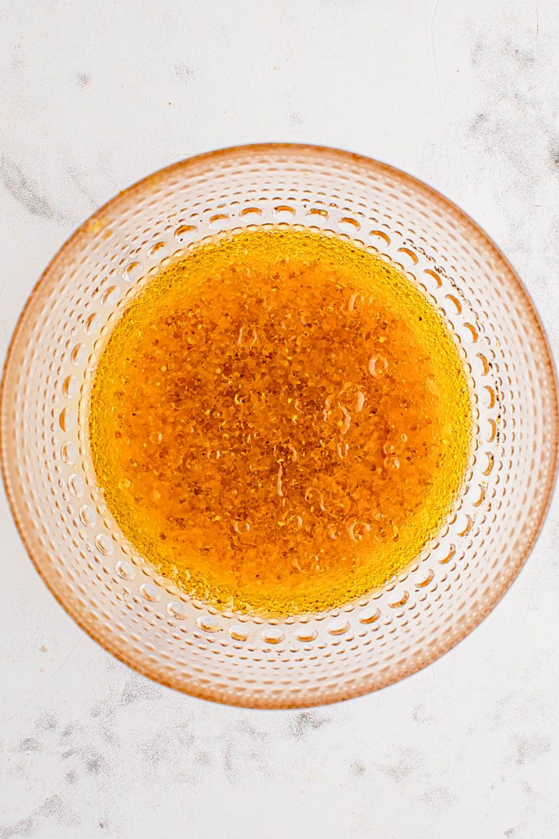 A glass bowl containing golden brown liquid with bubbles, viewed from above, on a light-colored surface.