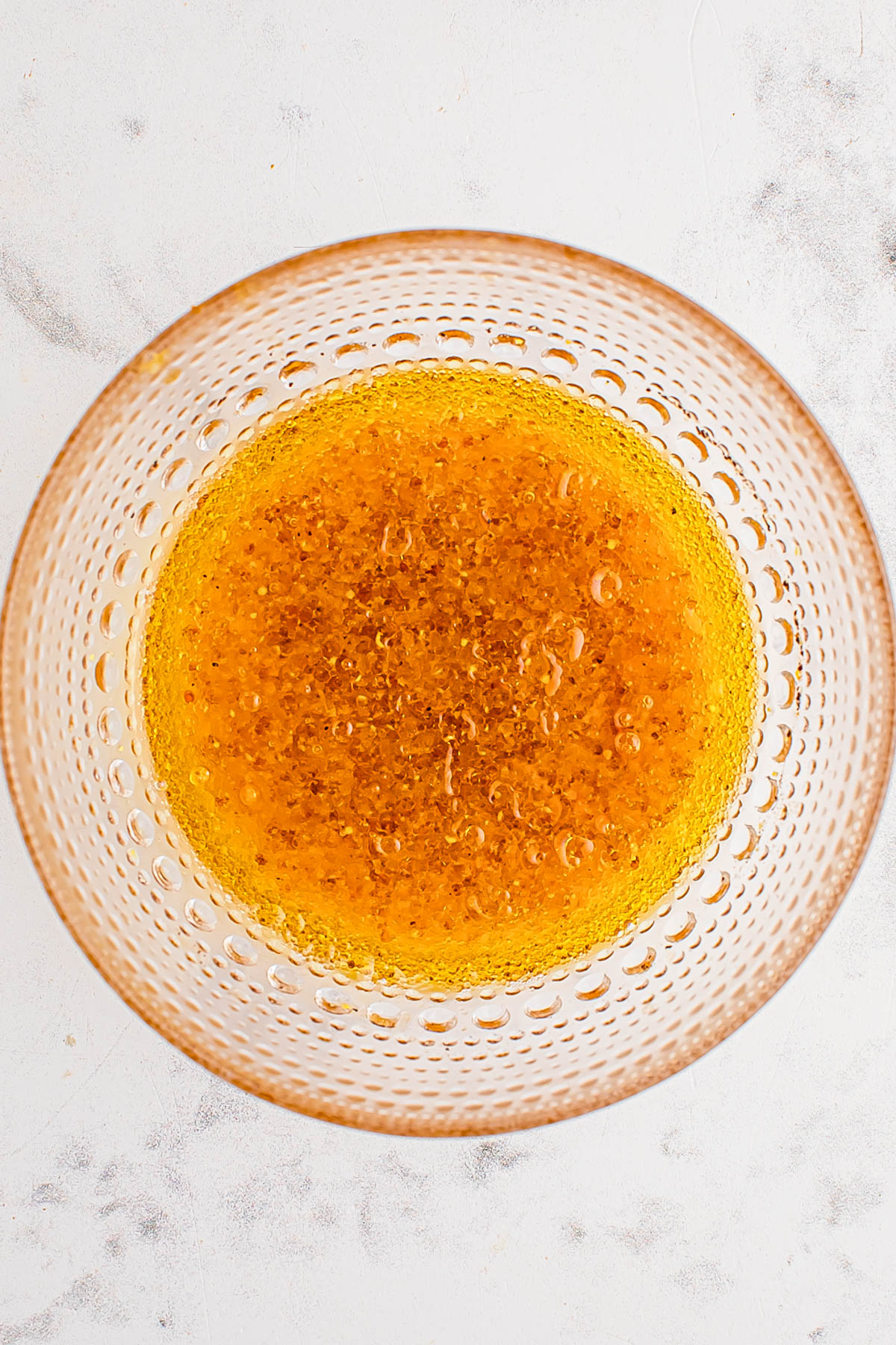A glass bowl containing golden brown liquid with bubbles, viewed from above, on a light-colored surface.