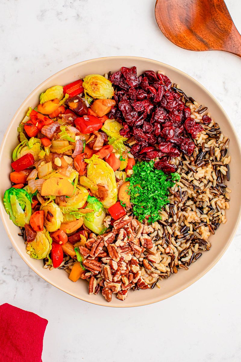 A bowl containing cooked wild rice, sautéed Brussels sprouts and vegetables, dried cranberries, chopped parsley, and pecans, arranged in separate sections on a white surface.