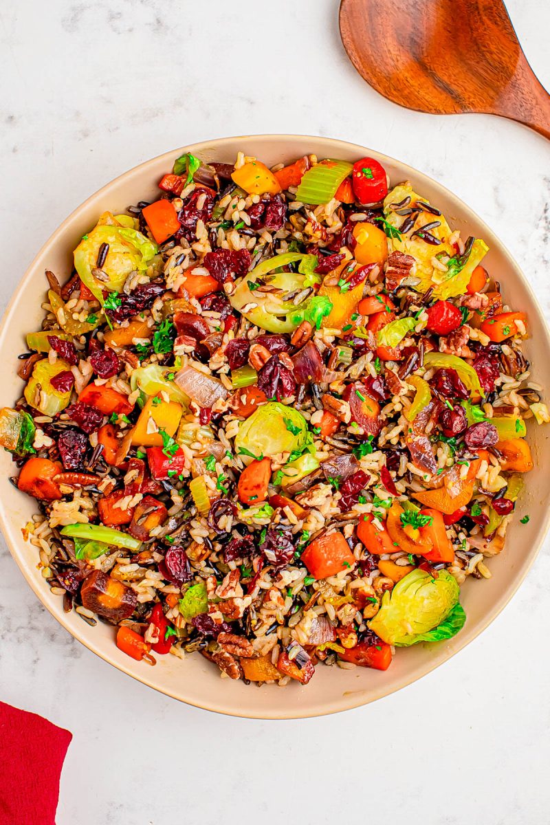 A bowl of mixed wild rice with Brussels sprouts, carrots, red bell peppers, dried cranberries, and fresh herbs on a white surface.