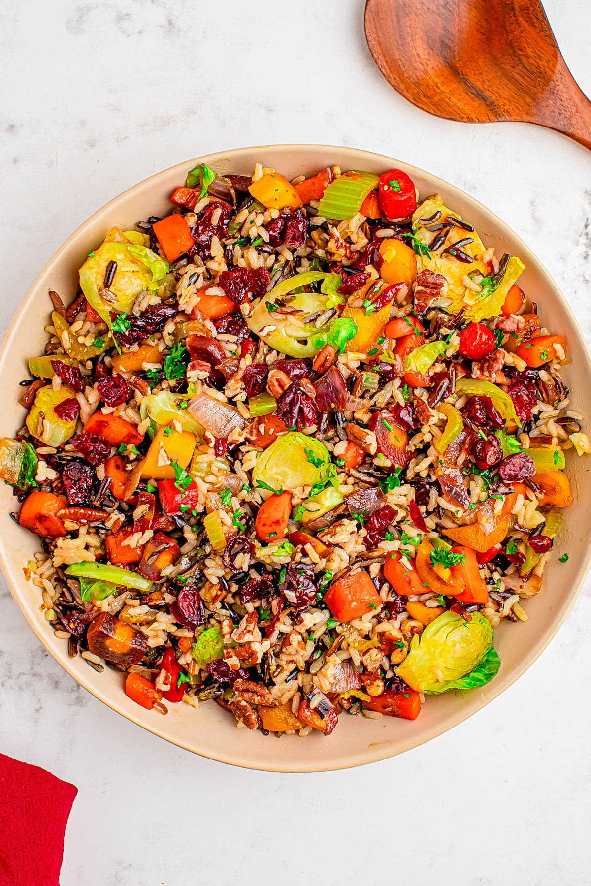 A bowl of mixed wild rice with Brussels sprouts, carrots, red bell peppers, dried cranberries, and fresh herbs on a white surface.