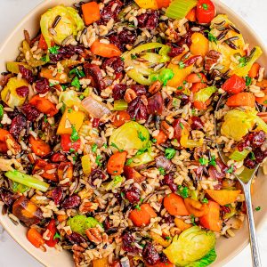 A bowl of mixed wild rice with Brussels sprouts, carrots, bell peppers, cranberries, and fresh herbs, with a spoon resting on the side.