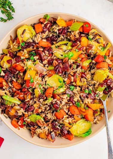 A bowl of mixed wild rice with Brussels sprouts, carrots, bell peppers, cranberries, and fresh herbs, with a spoon resting on the side.