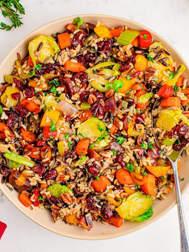 A bowl of mixed wild rice with Brussels sprouts, carrots, bell peppers, cranberries, and fresh herbs, with a spoon resting on the side.