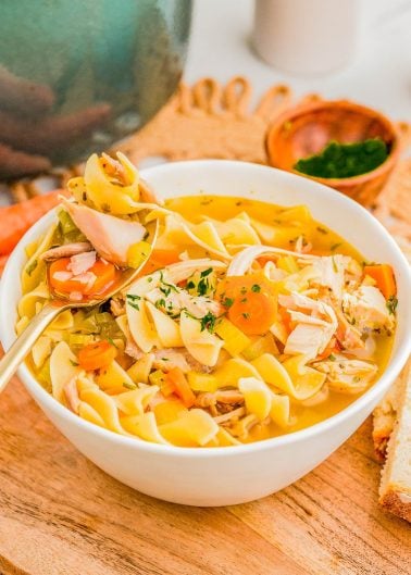 A bowl of chicken noodle soup with vegetables, garnished with herbs, sits on a wooden board next to a slice of bread.