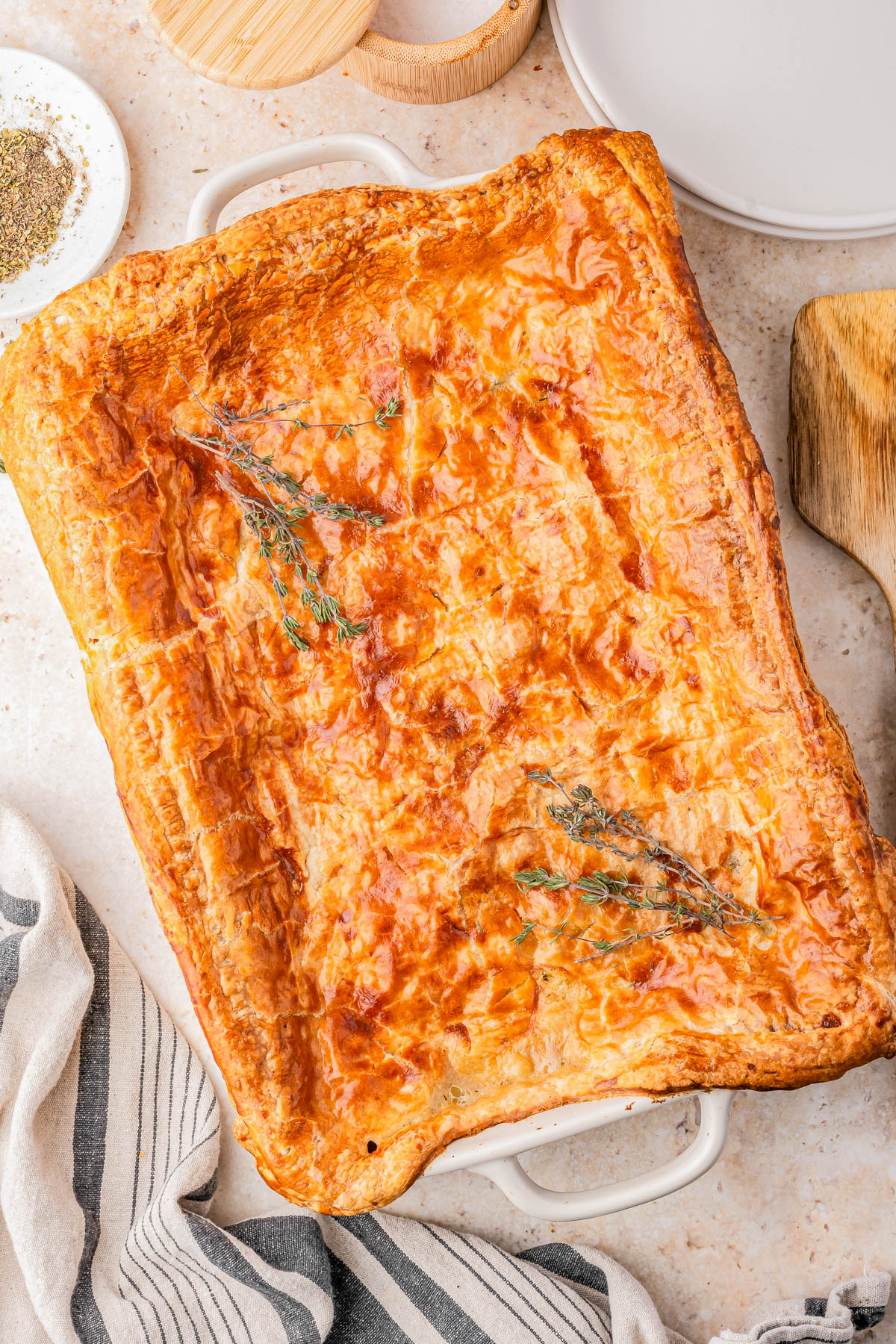 Golden-brown rectangular pie with a flaky crust, garnished with sprigs of herbs, in a white baking dish on a light countertop.