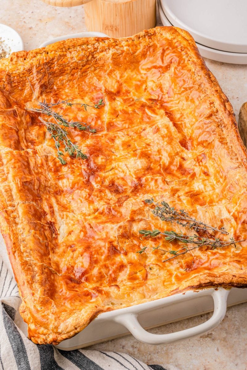 A golden-brown baked puff pastry pie in a rectangular dish, topped with sprigs of fresh herbs.
