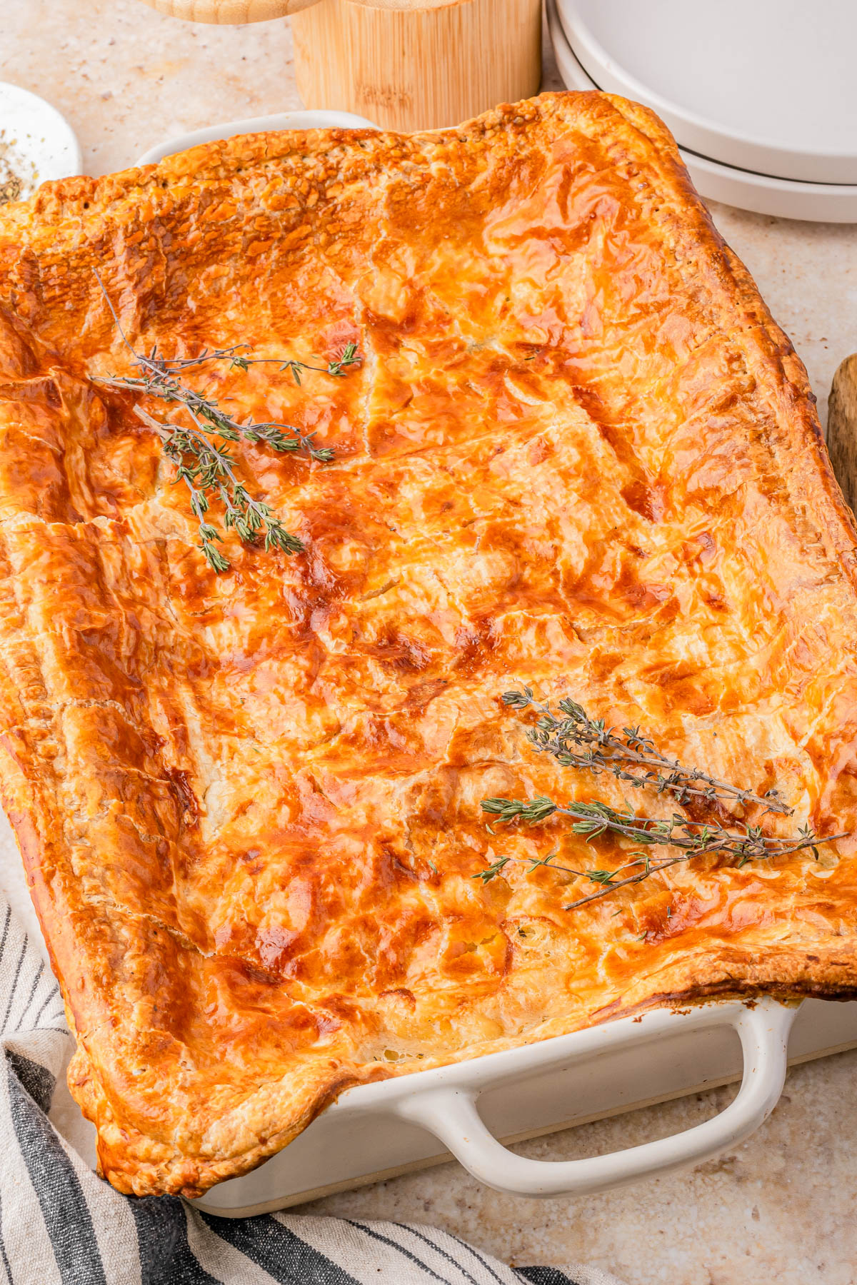 A golden-brown baked puff pastry pie in a rectangular dish, topped with sprigs of fresh herbs.
