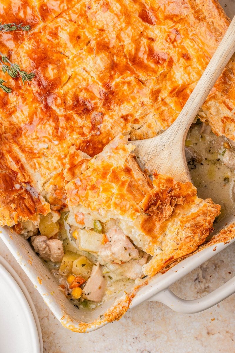 A golden-brown chicken pot pie with a flaky crust sits in a baking dish, a wooden spoon lifting a serving to reveal the filling of chicken, vegetables, and creamy sauce.