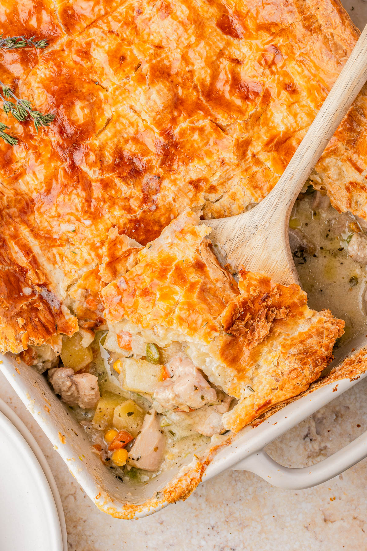 A golden-brown chicken pot pie with a flaky crust sits in a baking dish, a wooden spoon lifting a serving to reveal the filling of chicken, vegetables, and creamy sauce.