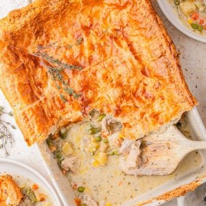 Golden-brown puff pastry-topped chicken pot pie in a baking dish, partially served with filling of chicken and mixed vegetables, surrounded by two plated servings.