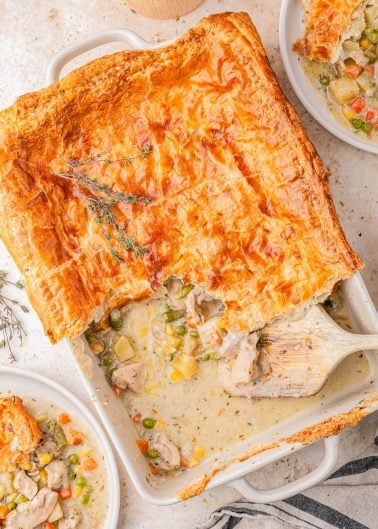 Golden-brown puff pastry-topped chicken pot pie in a baking dish, partially served with filling of chicken and mixed vegetables, surrounded by two plated servings.
