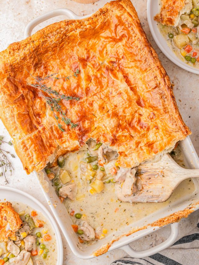 Golden-brown puff pastry-topped chicken pot pie in a baking dish, partially served with filling of chicken and mixed vegetables, surrounded by two plated servings.