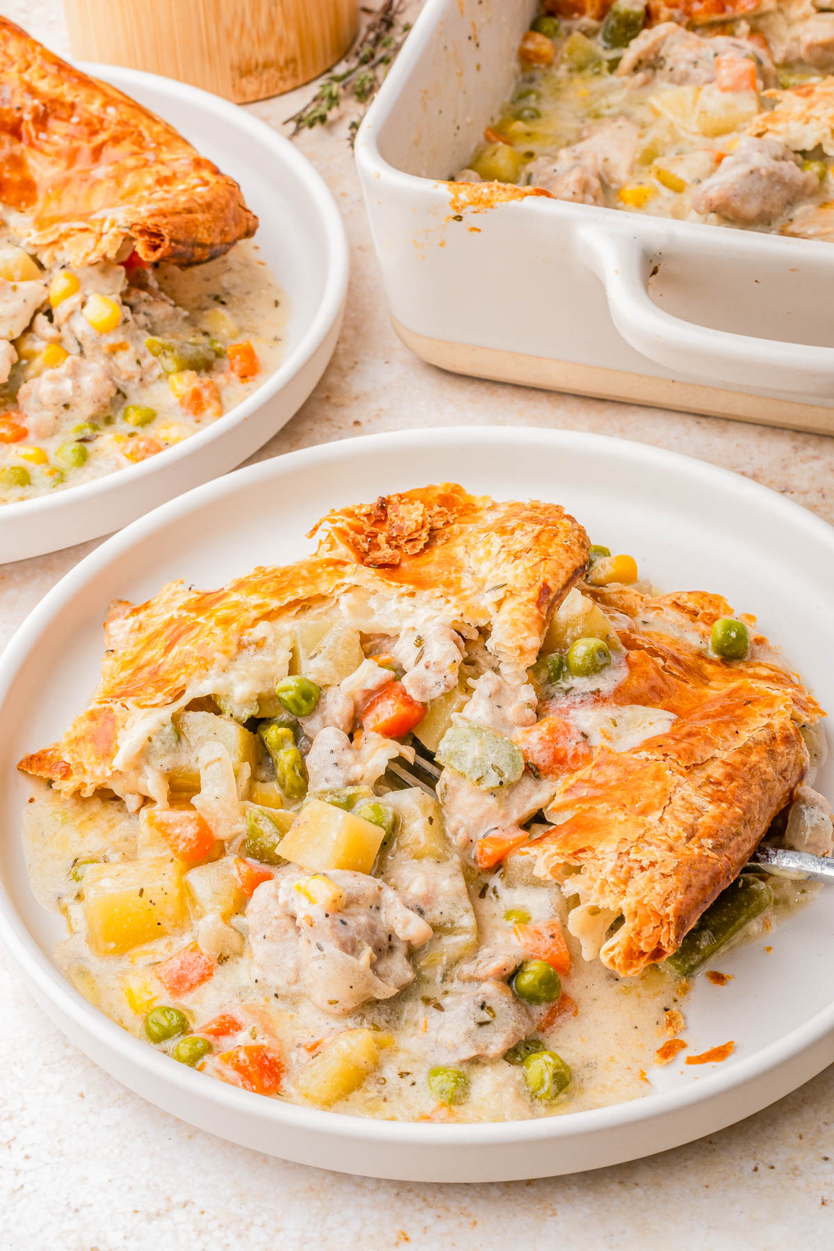 A serving of chicken pot pie with a golden, flaky crust on a white plate, showing mixed vegetables and creamy filling; a baking dish and another plate are in the background.