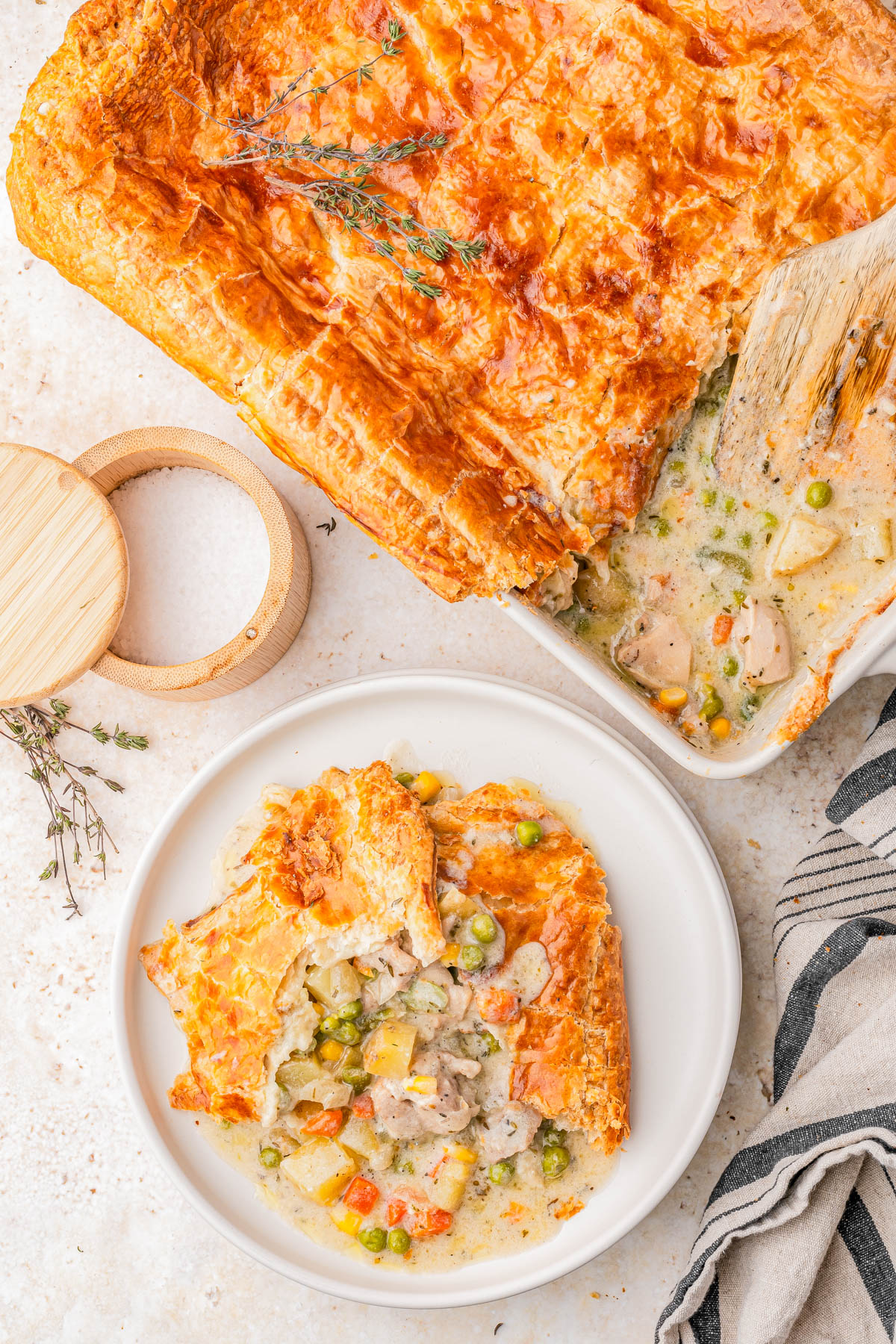 A serving of chicken pot pie with golden, flaky crust and mixed vegetables on a white plate, beside a baking dish containing the remaining pie.