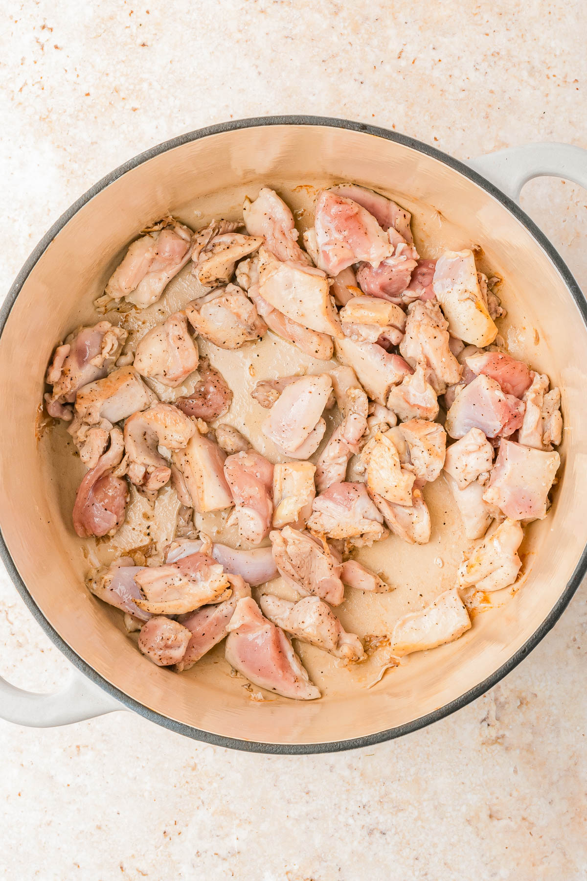 Raw, seasoned chicken pieces are spread out in a large, light-colored pot on a beige surface.