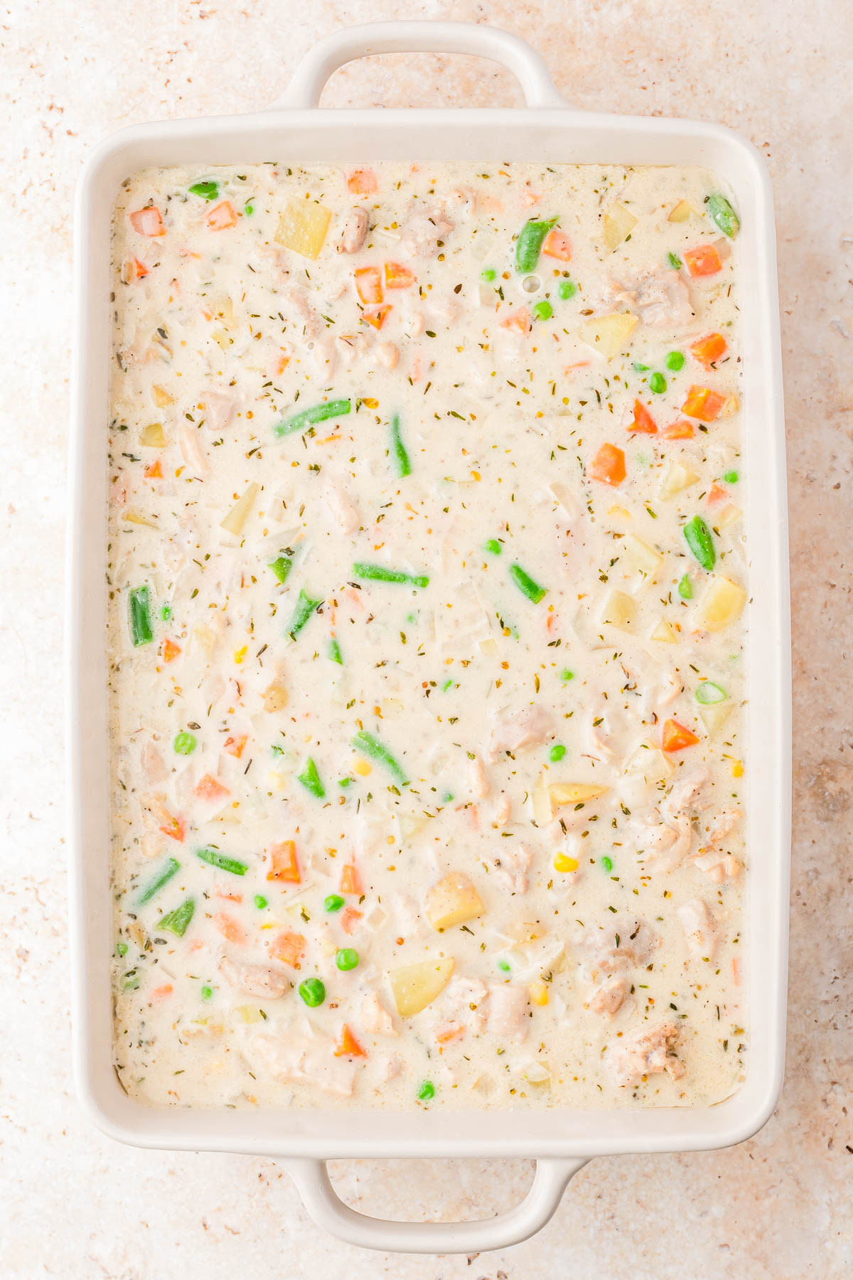A rectangular baking dish filled with a creamy mixture of diced chicken, mixed vegetables, and herbs in a white sauce.