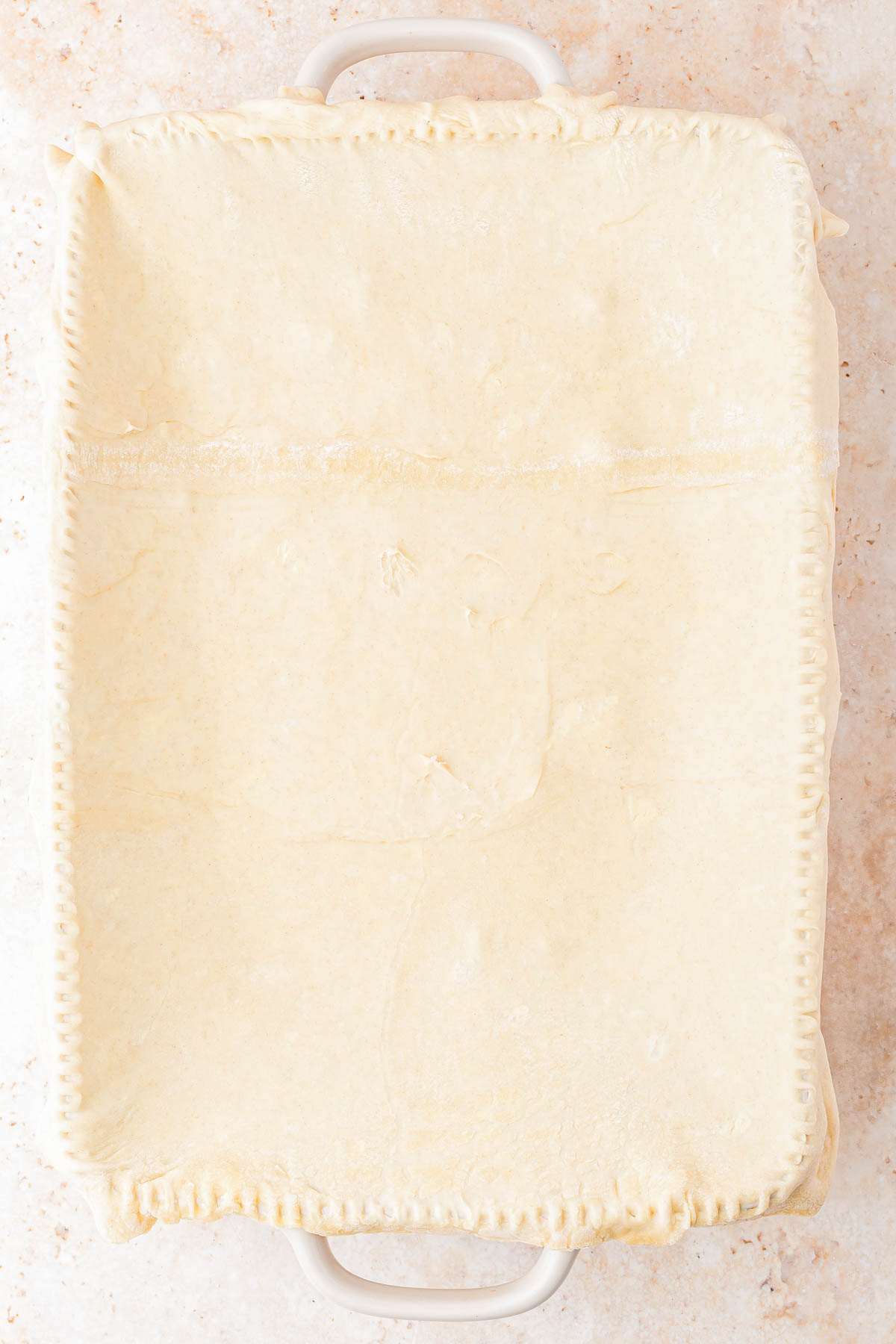 A rectangular baking dish covered with an uncooked sheet of pastry dough, edges sealed with a fork, on a light-colored surface.