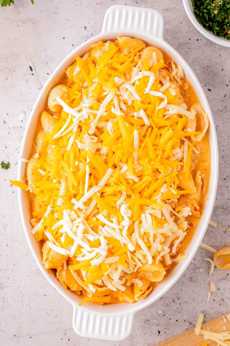 An oval white baking dish filled with macaroni and cheese, topped with shredded yellow and white cheese, on a light countertop.