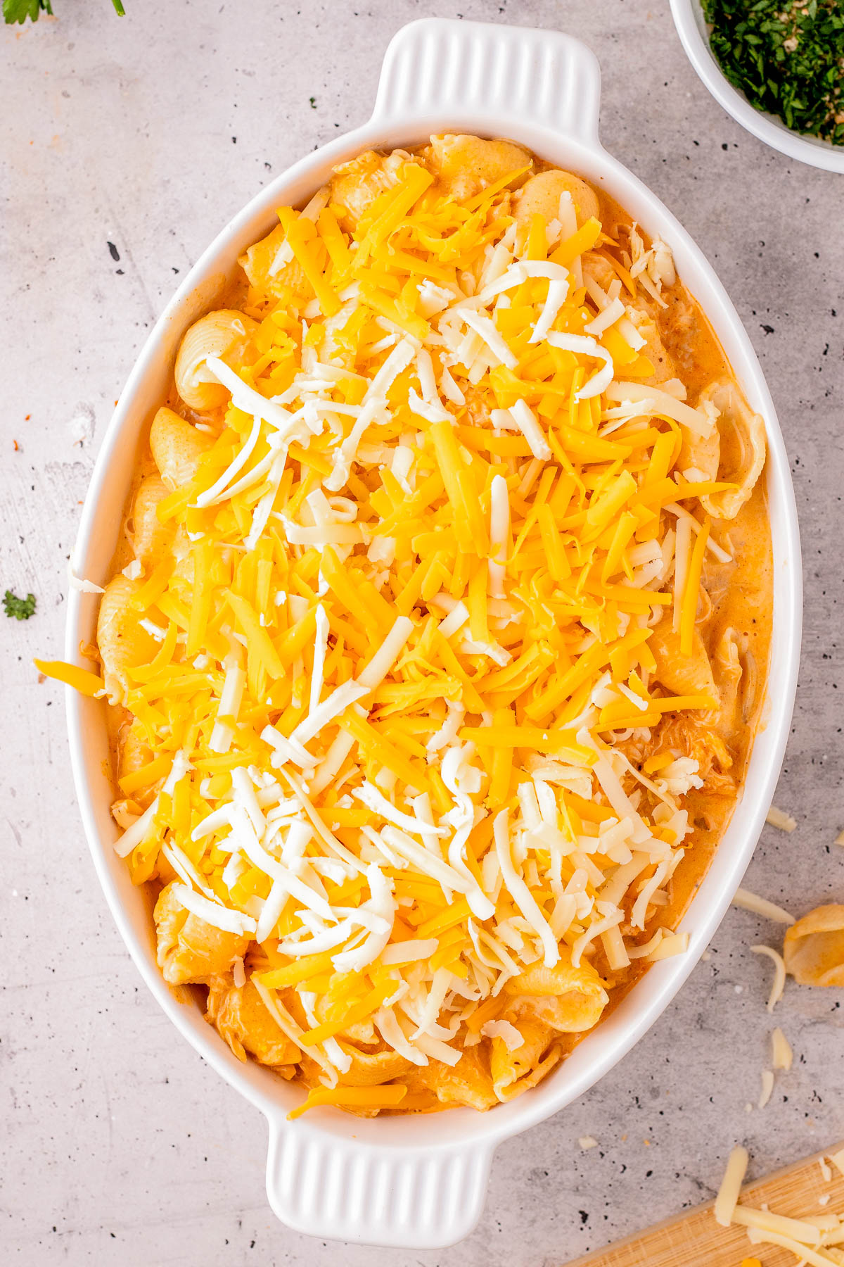 An oval white baking dish filled with macaroni and cheese, topped with shredded yellow and white cheese, on a light countertop.