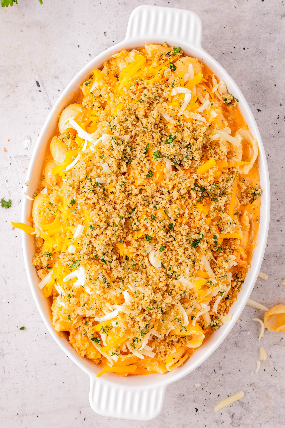 A white oval baking dish filled with a casserole topped with shredded cheese and a golden breadcrumb mixture on a light surface.