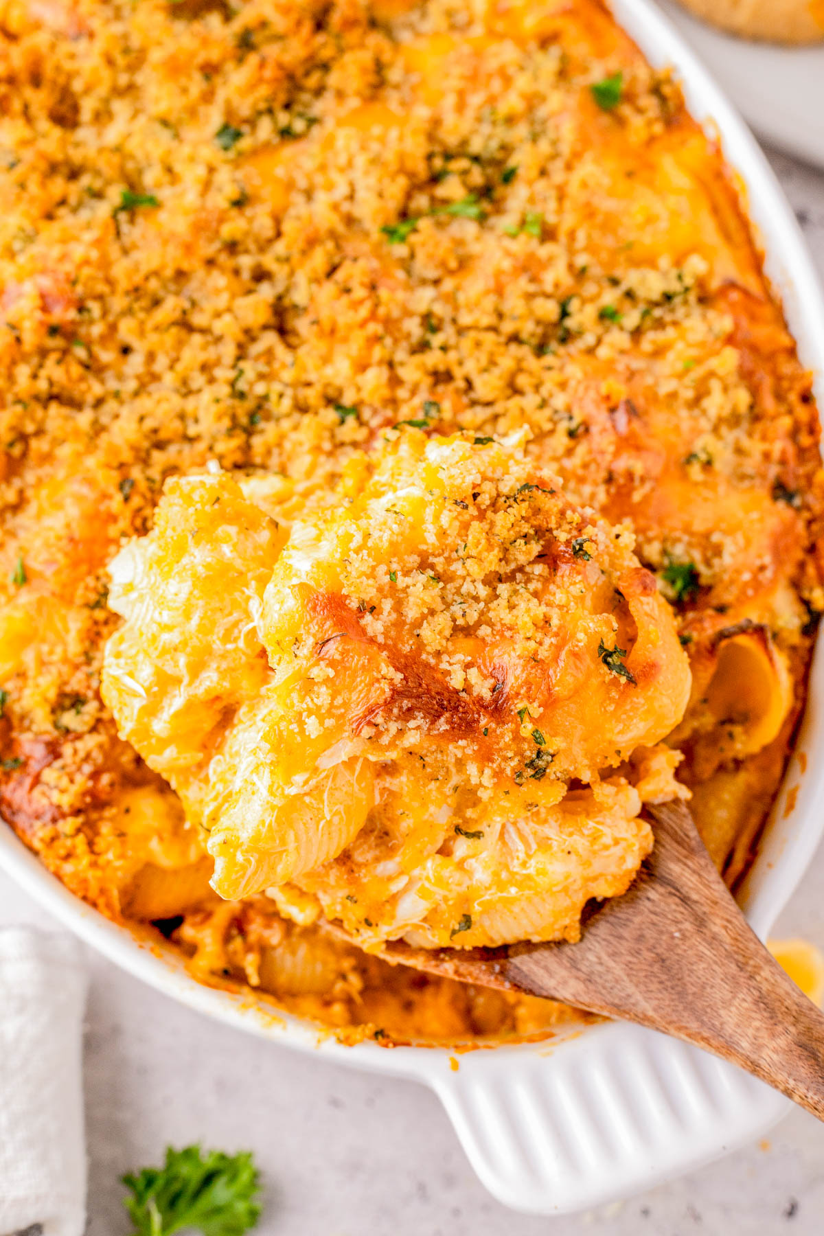 A wooden spoon lifts a serving of baked macaroni and cheese topped with golden breadcrumbs from a casserole dish.