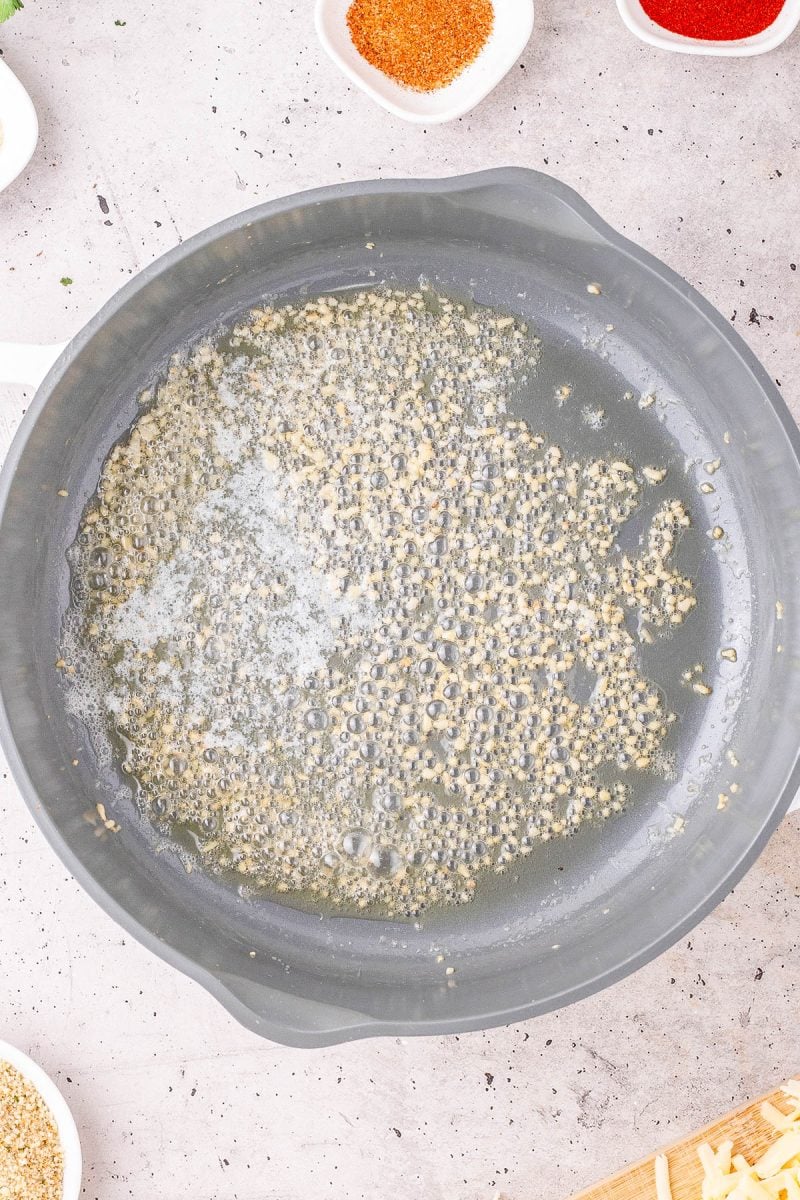 A frying pan with minced garlic sizzling in melted butter, surrounded by small bowls containing spices on a light countertop.
