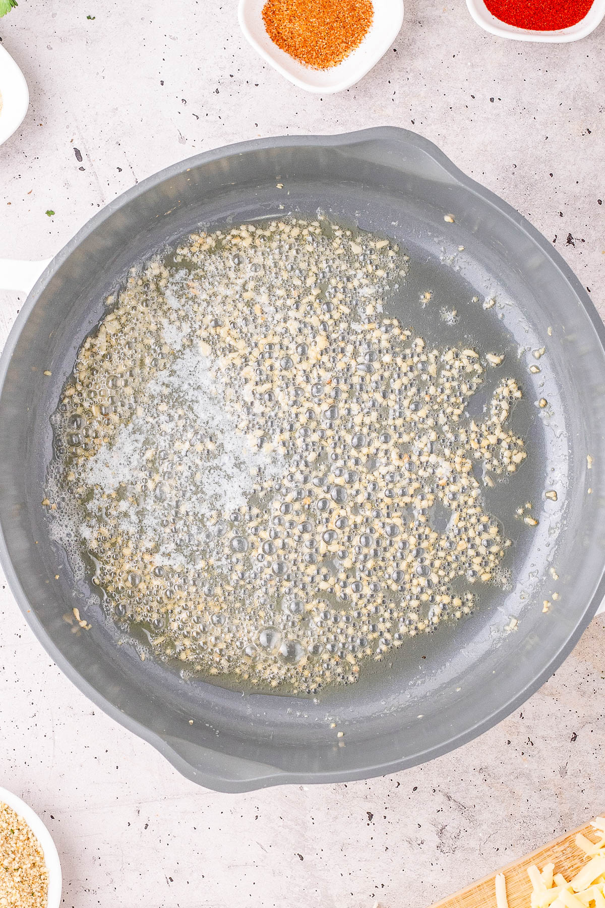 A frying pan with minced garlic sizzling in melted butter, surrounded by small bowls containing spices on a light countertop.