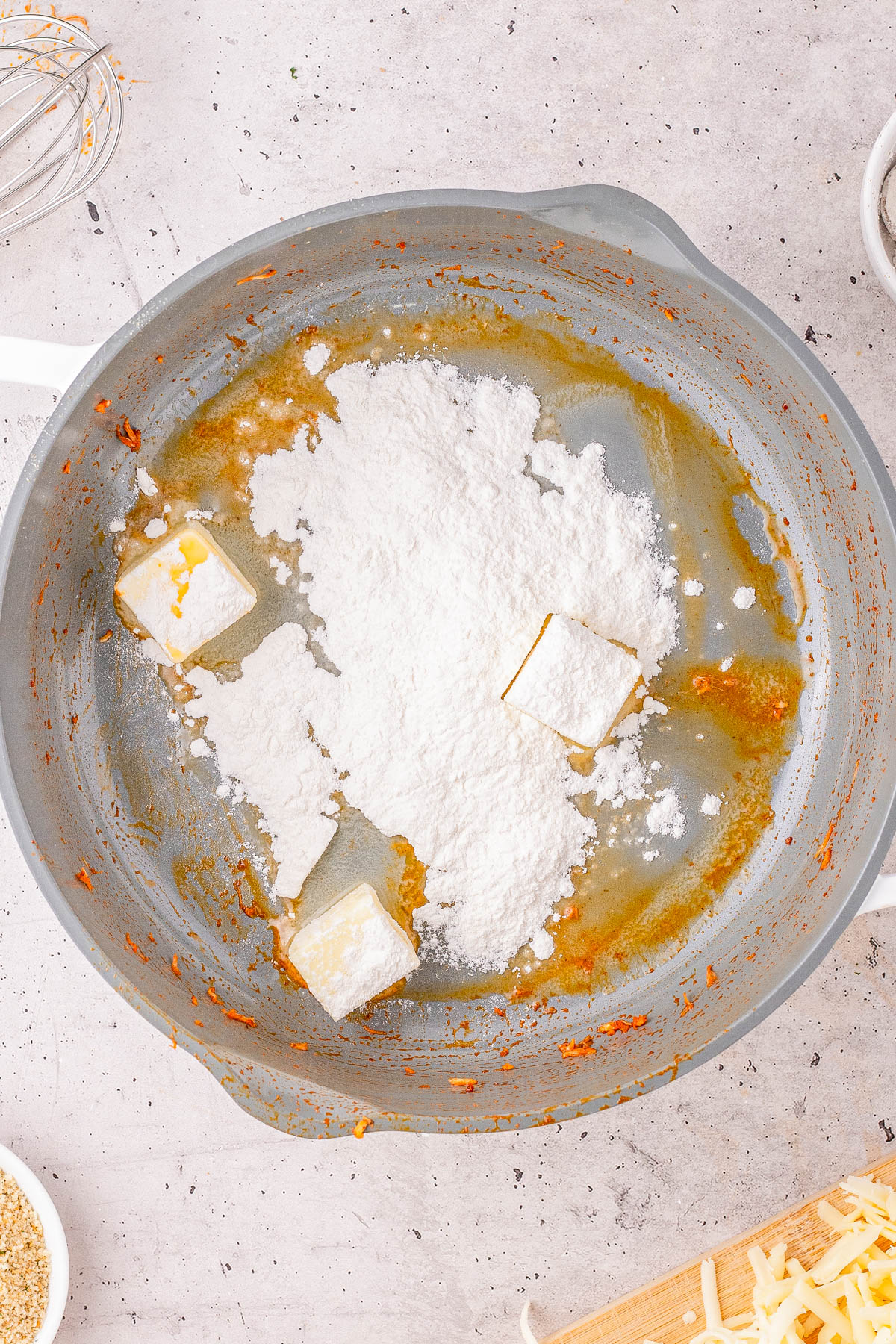 A saucepan with melted butter and flour being combined, surrounded by kitchen utensils and grated cheese on a light countertop.