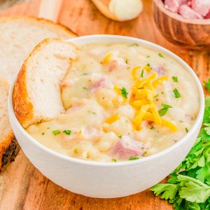 A bowl of creamy ham and potato soup garnished with shredded cheese and parsley, served with a slice of bread on a wooden board. Chopped ham and herbs are in the background.