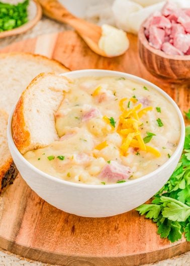 A bowl of creamy ham and potato soup garnished with shredded cheese and parsley, served with a slice of bread on a wooden board. Chopped ham and herbs are in the background.