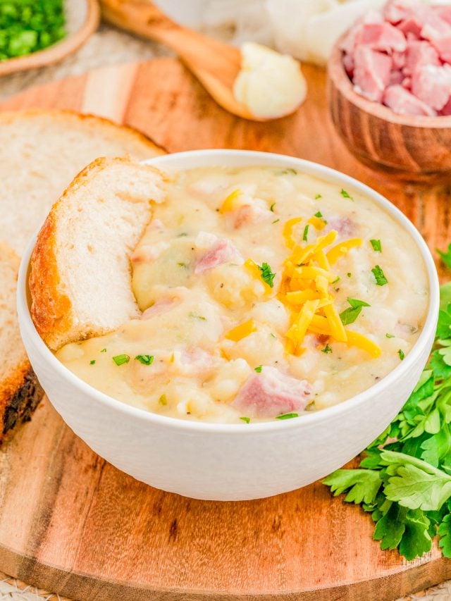 A bowl of creamy ham and potato soup garnished with shredded cheese and parsley, served with a slice of bread on a wooden board. Chopped ham and herbs are in the background.