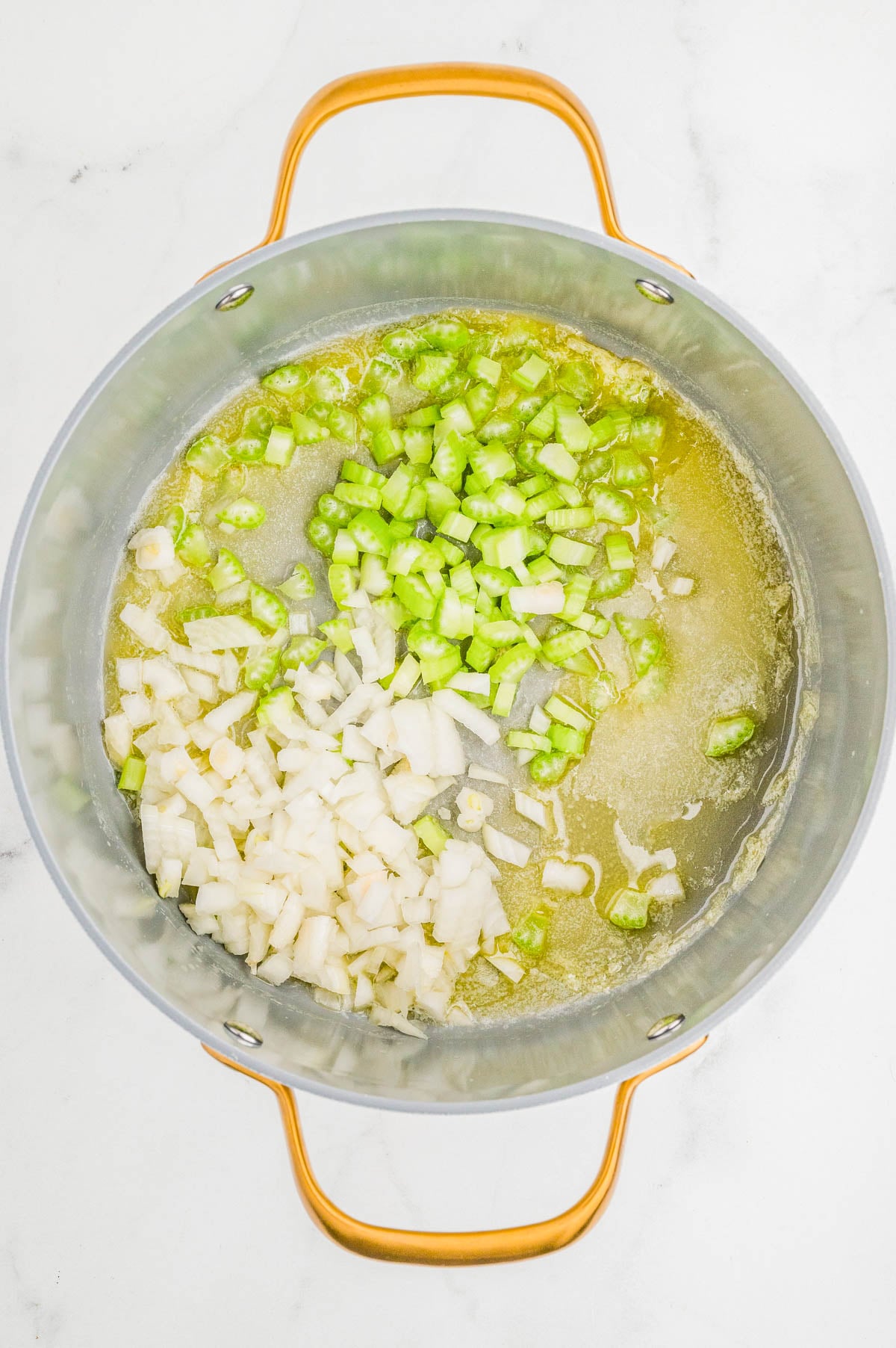 Chopped celery and onions cooking in melted butter in a large pot on a white surface.